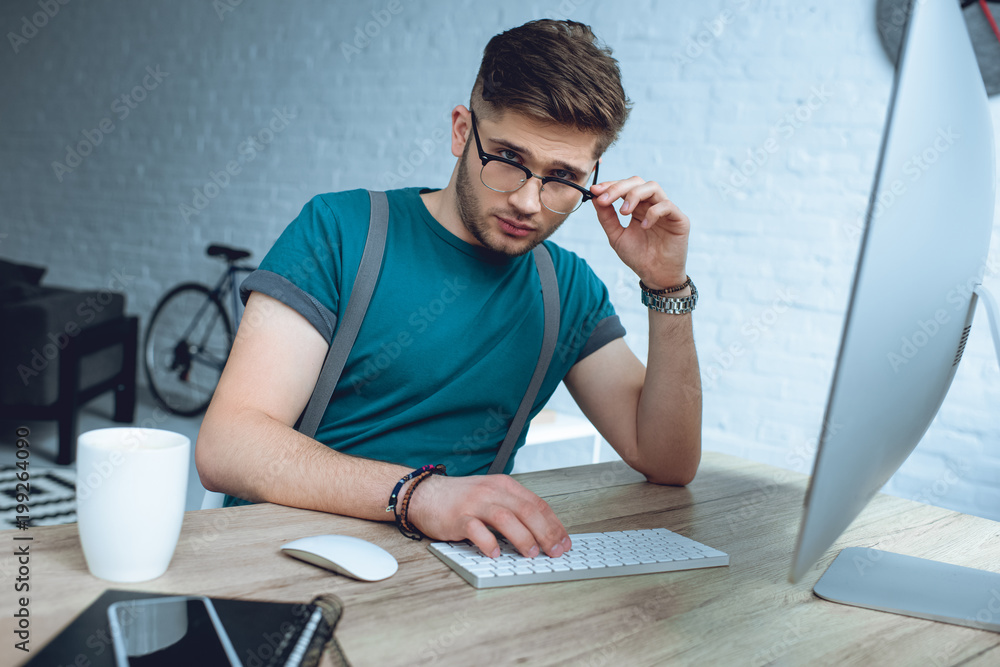 Poster handsome young freelancer adjusting eyeglasses and looking at camera while working with desktop comp