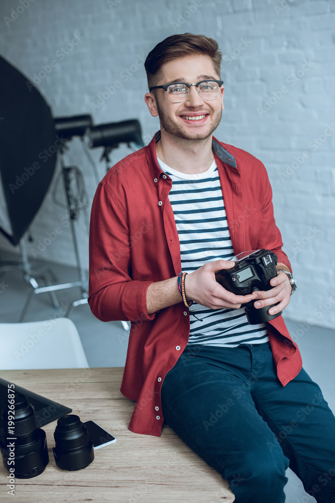 Poster smiling man holding camera and sitting on table with computer at home office
