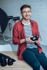 Smiling man holding camera and sitting on table with computer at home office