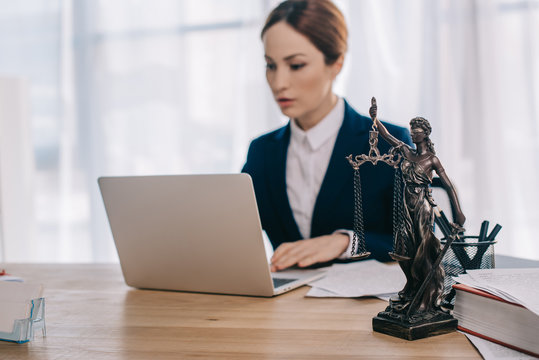 Selective Focus Of Female Lawyer In Suit Working On Laptop At Workplace With Femida In Office