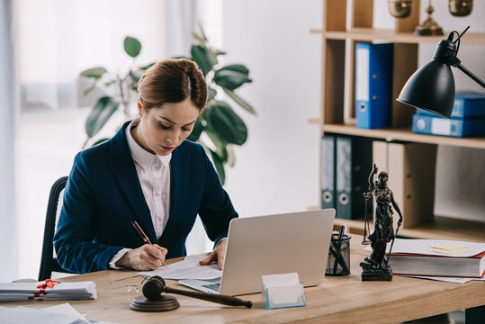 Female Lawyer In Suit At Workplace With Laptop, Gavel And Femida In Office