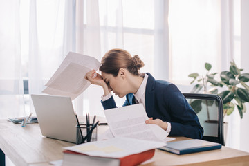 overworked businesswoman in suit with papers in hands at workplace in office