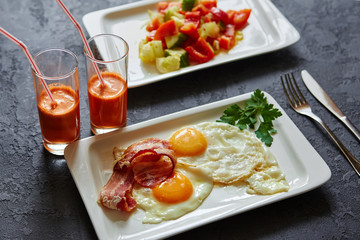 Breakfast from fried egg, bacon, vegetable salad and carrot juice. On a dark stone background