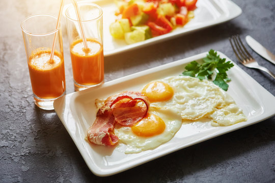 Breakfast from fried egg, bacon, vegetable salad and carrot juice. On a dark stone background