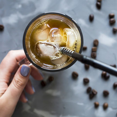 A female hand holds a black cold coffee with ice and milk on a gray background among the coffee beans. Summer cooling drink in a glass cup. Top view, flat lay