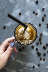 A female hand holds a black cold coffee with ice and milk on a gray background among the coffee beans. Summer cooling drink in a glass cup. Top view, flat lay