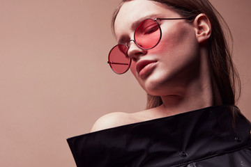 Warm colored fashion portrait of beautiful young woman in round red sunglasses and grey latex jacket posing in studio - 199284842