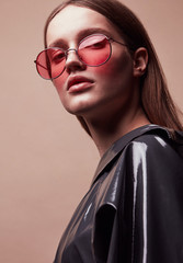 Fashion portrait of beautiful young woman in round red sunglasses and grey latex jacket posing in studio - 199284843