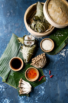 Asian rice piramidal steamed dumplings from rice tapioca flour with meat filling in banana leaves served in bamboo steamer. Ingredients and sauces above over blue texture background. Top view, space.
