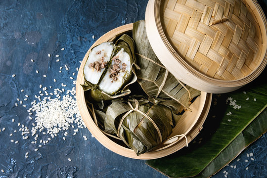 Asian rice piramidal steamed dumplings from rice tapioca flour with meat filling in banana leaves served in bamboo steamer with rice above over blue texture background. Top view, space.