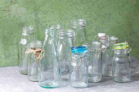 Variety of different shape empty opened glass bottles with and without lids standing on grey table with green wall as background.