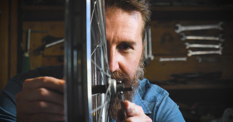 Well-groomed handsome bearded master hipster, specialist in bicycles, repairing a bicycle in his workshop, wheels, frame, spokes, the background of tools. Concept: pro bike, cycle passion, lifestyle.