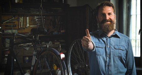 Obraz na płótnie Canvas Well-groomed handsome bearded master hipster, specialist in bicycles, repairing a bicycle in his workshop, wheels, frame, spokes, the background of tools. Concept: pro bike, cycle passion, lifestyle.