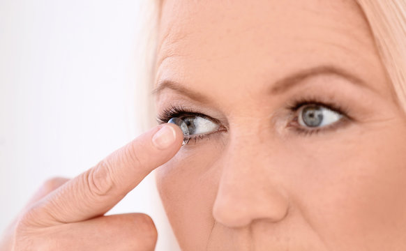 Senior woman putting contact lens in her eye on light background