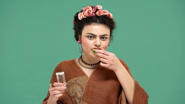 Portrait of charming happy woman with thick eyebrows and red lips drinking tequila with lime, isolated over green background. Stylization concept