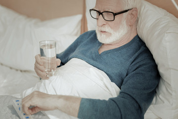 Illness and me. Unhappy serious bespectacled man lying on the bed taking pills and holding a glass of water.