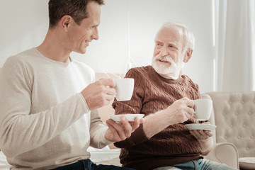 Warm smiles. Satisfied sociable happy men spending time in the bright room smiling and drinking tea.