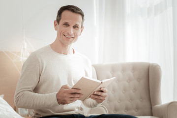 Interesting to know. Satisfied smart pleasant man sitting in the bright room holding a book and smiling.