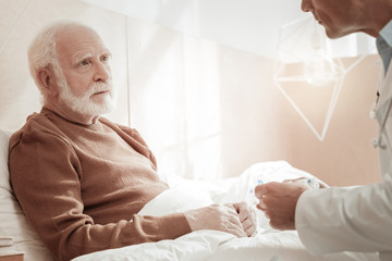 New hope. Pleasant interested aged man sitting on the bed looking at a doctor and listening.