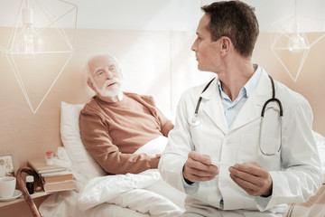 Fototapeta premium What is the diagnosis. Calm skilled handsome medic sitting on the bed holding the thermometer and turning to his patient.