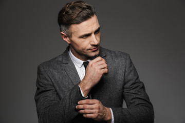 Fashion image of masculine man dressed in business suit looking aside, while fasten cufflink or button on sleeve of jacket, isolated over gray background