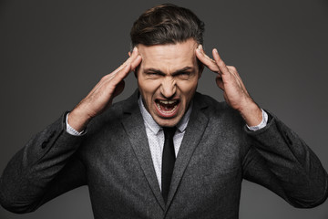 Photo of tired overworked businessman wearing formal suit squeezing his temples and screaming, isolated over gray background