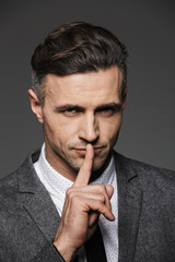 Photo of trendy man dressed in businesslike suit looking on camera and showing silence gesture with finger on lips, isolated over gray background