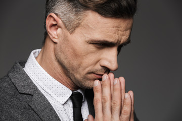 Fototapeta premium Photo closeup of thoughtful calm man wearing classic costume and tie keeping palms together in praying pose, isolated over gray background