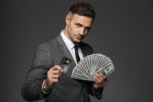 Photo of rich man 30s in business suit holding fan of dollar cash and digital money on credit card, isolated over gray background