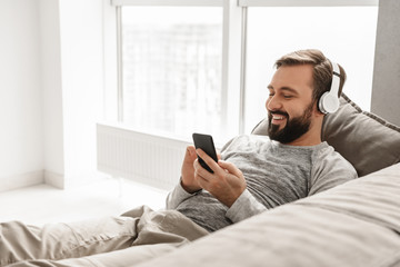Bearded man 30s in basic clothing lying on sofa at home, and listening to music on black cell phone wearing wireless headphones