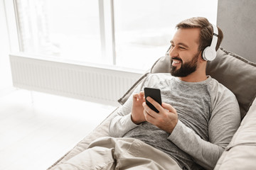 Joyful man with european appearance in basic clothing lying on sofa in house, and listening to music using black cell phone wearing wireless headphones
