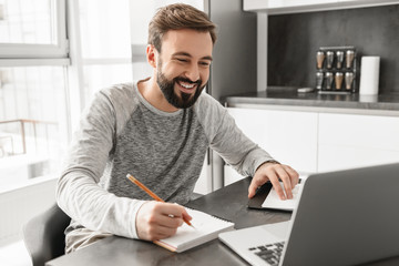 Photo of successful mature man 30s in casual clothing smiling and writing down notes on paper and...