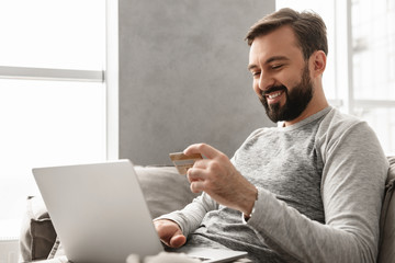 Image of caucasian unshaved man 30s in casual wear sitting on sofa in living room, and paying...