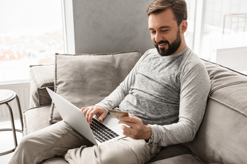 Pleased modern man 30s in casual wear sitting on sofa in living room, and doing paying transaction with credit card and notebook