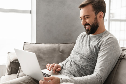 Photo of happy bearded man 30s in casual clothing working on laptop at modern apartment