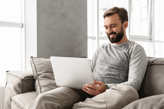 Photo of pleased adult man 30s in casual clothing typing on laptop, while working in cozy home