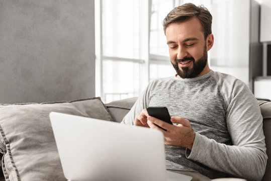 Photo of caucasian man 30s in casual wear using smartphone, while working on laptop in living room