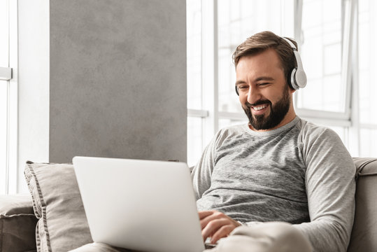 Photo of content adult man 30s in casual clothing resting at home, and listening to music via headphones on laptop