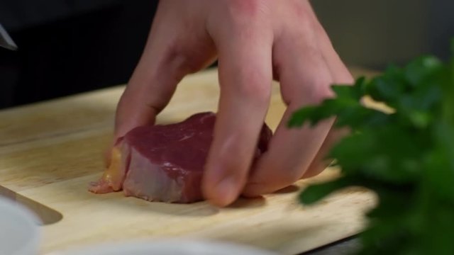 Hands of male cook cutting piece of raw meat on wooden cutting board and putting it into bowl, tracking left