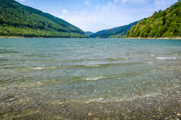 Mountain lake shore with small waves in foreground, cloudy sky autumn day and distant green hills at horizon in the background