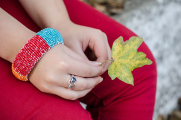 Girl in red pants wearing a big bracelet on her wrist and beautiful heart ring on her finger is holding a colorful autumn leaf in her hands