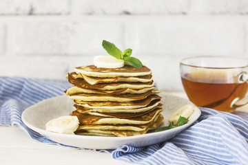 fresh baked pancakes with banana in white plate and cup of tea on blue linen on white background