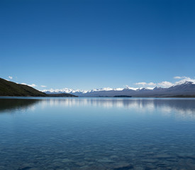 Fototapeta na wymiar Imagen de un paisaje azul minimalista con montañas nevadas distantes y un lago enorme y claro. Las montañas se reflejan en el agua. Y hay muchas rocas y piedras en el fondo del lago.