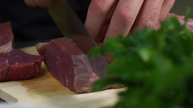 Close up hands of male cook cutting raw meat into pieces on wooden cutting board, tracking left