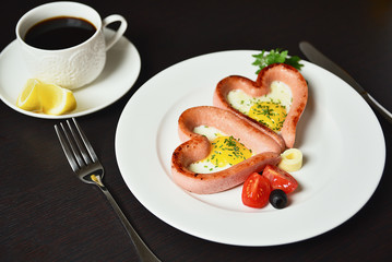 Scrambled eggs in the form of heart on a white plate with sausages, tomatoes, greens and coffee with lemon.