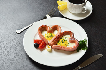 Scrambled eggs in the form of heart on a white plate with sausages, tomatoes, greens and coffee with lemon.