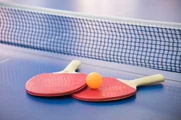 Orange ball for table tennis and two rackets of red and black color on a blue table with a grid
