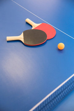 Orange ball for table tennis and two rackets of red and black color on a blue table with a grid