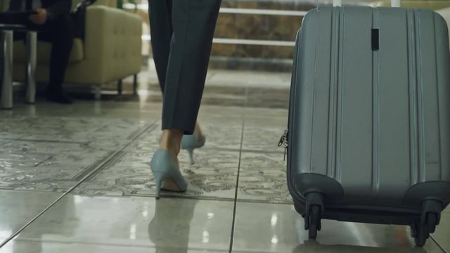 Dolly shot of legs of businesswoman walking through hotel lobby pulling luggage and stop at reception desk. Business, travel and people concept