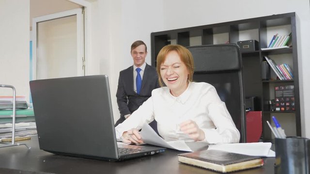 Two office workers sit at the desk, a woman is working on the computer, a man is near. They are laughing. 4k